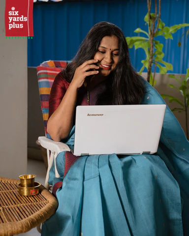 Woman in a blue saree smiling while using a laptop indoors, seated on a white chair with a colorful cushion.