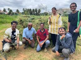 A group of six people smiling and posing on a farm field with greenery and buildings in the background.