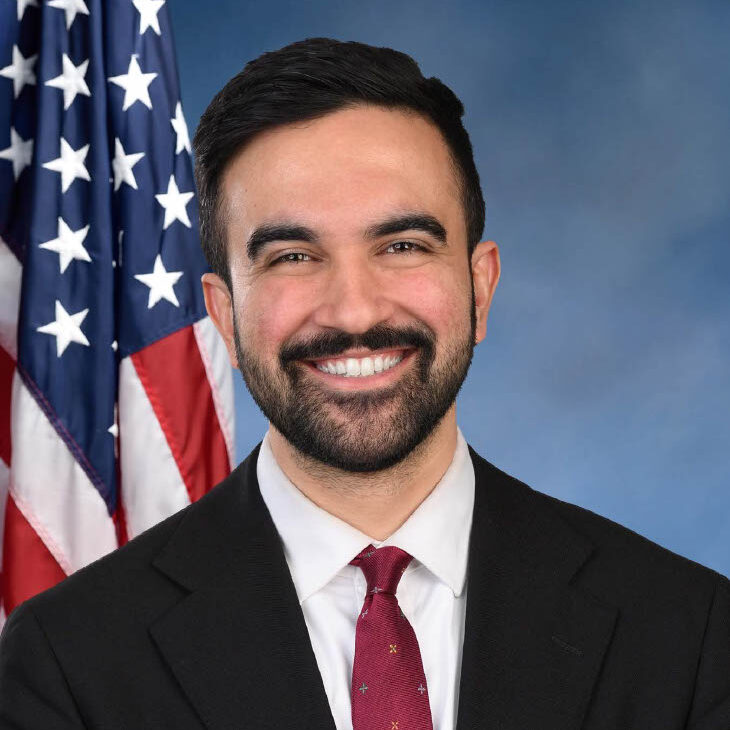 Zohran Mamdani, American politician and New York Assembly member, smiling in a formal portrait with the U.S. flag in the background.