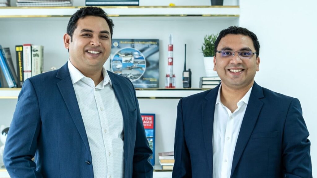 Two men in formal suits smiling in an office setting with books and models in the background.